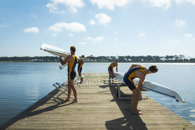 Side view of a rowing team of four Caucasian men preparing rowing boats before rowing standing on a jetty on the river on a sunny day — Stock Photo