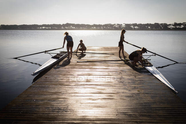 Vista lateral de uma equipe de remo de quatro mulheres caucasianas treinando no rio, em um molhe ao nascer do sol preparando dois barcos antes de remar — Fotografia de Stock