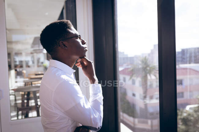 Un hombre de negocios afroamericano, con una camisa blanca, trabajando en una oficina moderna, mirando por la ventana, tocándose la barbilla y pensando: - foto de stock