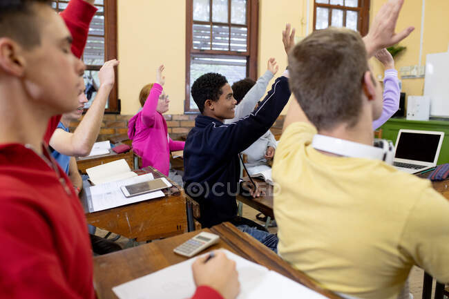 Vista lateral de un grupo multiétnico de adolescentes de secundaria en un aula escolar sentados en escritorios, todos levantando las manos para responder a una pregunta - foto de stock