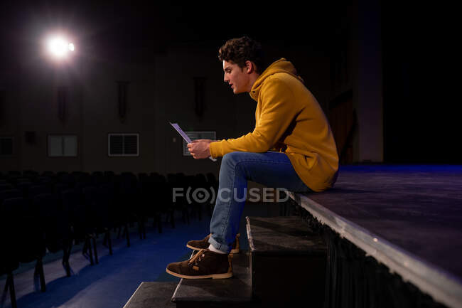 Seitenansicht eines kaukasischen Teenagers, der am Bühnenrand sitzt und in einem leeren Schultheater während der Proben für eine Aufführung ein Drehbuch hält — Stockfoto
