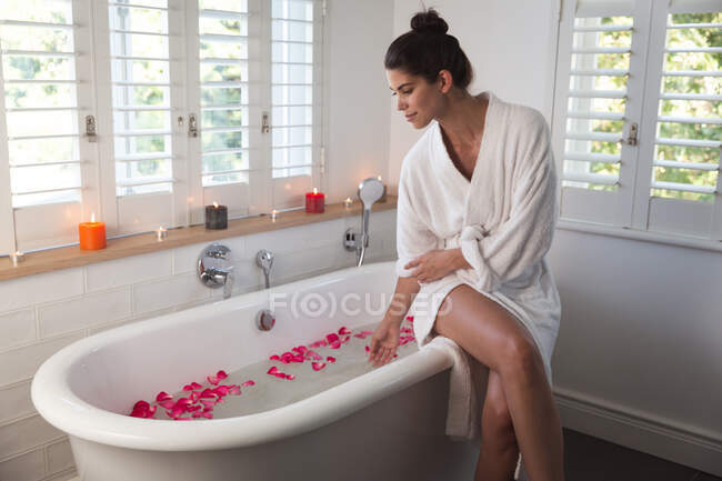 Femme de race mixte passant du temps à la maison, assis sur la baignoire bain de course dans la salle de bain. Auto-isolement et distanciation sociale en quarantaine pendant l'épidémie de coronavirus covid 19. — Photo de stock