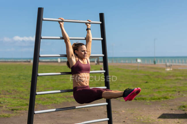 Vue latérale d'une femme sportive caucasienne aux longs cheveux foncés faisant de l'exercice dans une salle de gym extérieure au bord de la mer pendant la journée, suspendue à un cadre d'exercice tirant ses jambes droites vers le haut. — Photo de stock