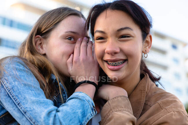 Vista frontal de cerca de un caucásico y una mezcla de chicas de raza disfrutando del tiempo juntos en un día soleado, sonriendo y susurrando, chica con frenillos. - foto de stock