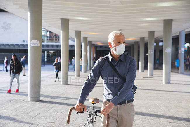 Hombre caucásico mayor fuera y alrededor de las calles de la ciudad durante el día, con una máscara facial contra el coronavirus, covid 19, ruedas de una bicicleta. - foto de stock