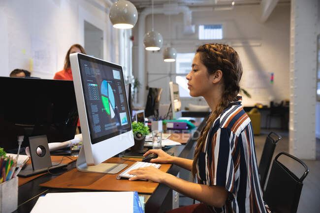 Frauen mit gemischter Rasse arbeiten kreativ in einem lässigen modernen Büro, sitzen an einem Tisch und benutzen einen Computer mit Kollegen, die im Hintergrund arbeiten. — Stockfoto