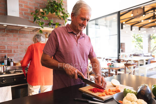 Glücklicher Rentner aus dem Kaukasus, der an einem sonnigen Tag zu Hause in der Küche steht, an der Arbeitsplatte Gemüse auf einem Schneidebrett schneidet und lächelt, während seine Partnerin im Hintergrund am Herd das Essen zubereitet — Stockfoto