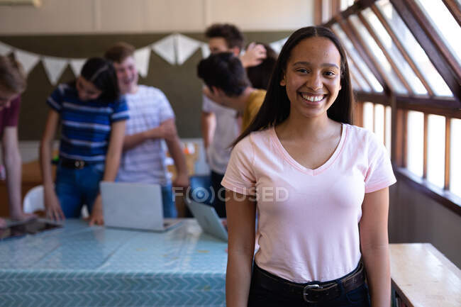 Portrait d'une adolescente métisse aux longs cheveux foncés et aux yeux bruns debout dans une classe d'école souriant à la caméra, avec des camarades de classe parlant en arrière-plan — Photo de stock