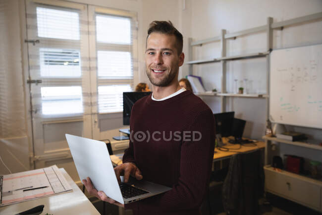 Porträt eines kaukasischen männlichen Unternehmens, das kreativ in einem lässigen modernen Büro arbeitet, Laptop hält und benutzt, in die Kamera schaut und lächelt, während Kollegen im Hintergrund arbeiten — Stockfoto