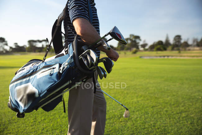 Vista lateral parte média do homem em um campo de golfe em um dia ensolarado com céu azul, andando e carregando um saco de golfe — Fotografia de Stock