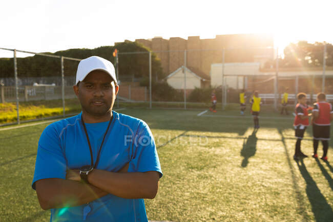 Портрет мужчины-тренера смешанной расы, стоящего на игровом поле под солнцем с скрещенными руками, смотрящего в камеру во время футбольной тренировки, с многонациональной группой мальчиков-футболистов на заднем плане, подсветка — стоковое фото