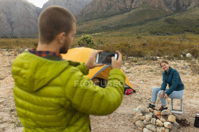 Side view of a Caucasian couple having a good time on a trip to the mountains, a woman is sitting by a campfire, cooking sausages on the sticks, while a man is standing and doing pictures of her — Stock Photo