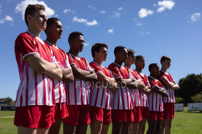 Vista lateral de un grupo de jugadores de rugby masculinos multiétnicos adolescentes con franja de equipo roja y blanca, de pie en un campo de juego con los brazos cruzados mirando hacia otro lado con un cielo azul en el fondo - foto de stock