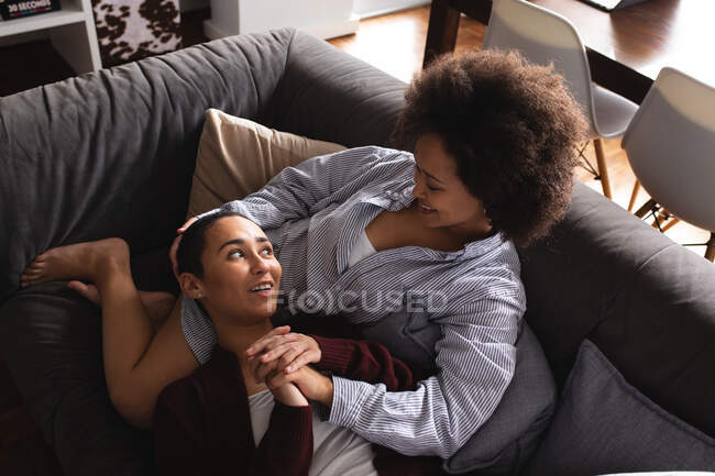 Високий кут зору змішаної раси жіноча пара розслабляється вдома у вітальні на дивані разом вранці, одна жінка лежить з головою на колінах свого партнера, тримаючись за руки і дивлячись один на одного посміхаючись — стокове фото