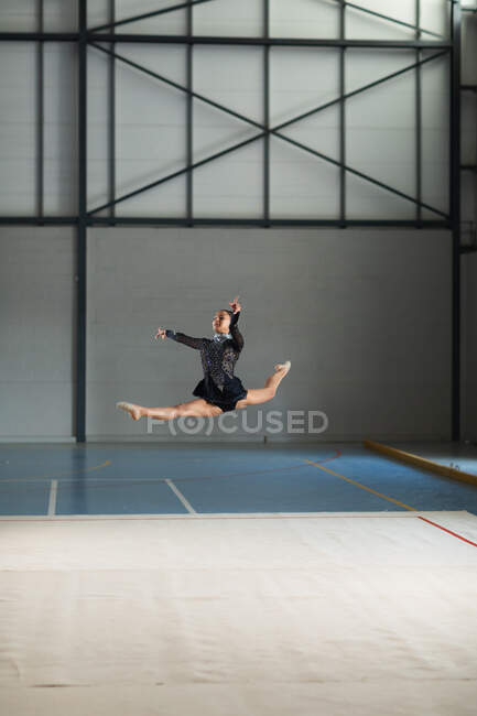 Вид спереду підлітків змішаної раси гімнастка, яка виступає в спортзалі, стрибає і робить розкол, одягнена в чорний леопард . — стокове фото