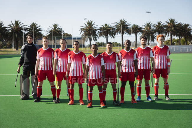 Front view of a multi-ethnic team of teenage male field hockey players preparing before a game, standing on the pitch in a row, wearing their team strip, holding their hockey sticks, looking to camera on a sunny day — Stock Photo
