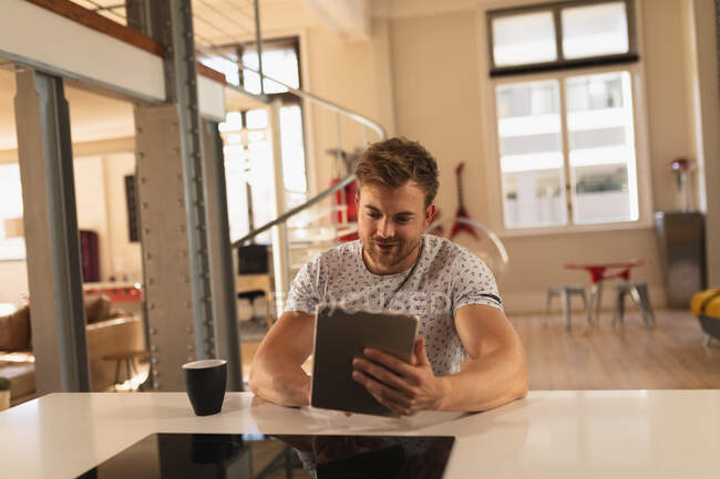 Front view of a young Caucasian man spending time at home, sitting in the kitchen with a cup of beverage, using his tablet. — Stock Photo