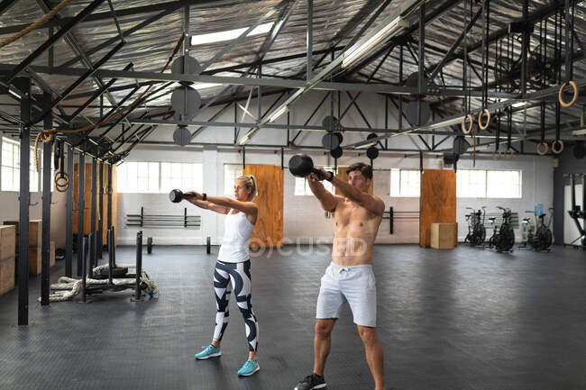 Front view of a shirtless athletic Caucasian man and woman wearing sports clothes cross training at a gym, weight training with kettlebells, standing, holding the kettlebells in both hands and lifting them to shoulder height — Stock Photo