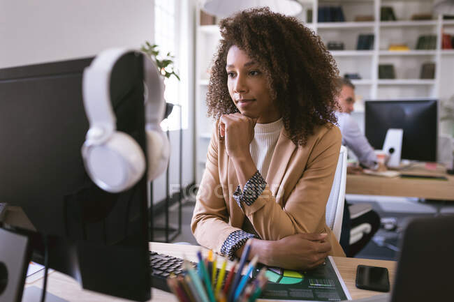 Una mujer de negocios de raza mixta que trabaja en una oficina moderna, sentada en un escritorio y usando una computadora, con sus colegas de negocios trabajando en segundo plano - foto de stock