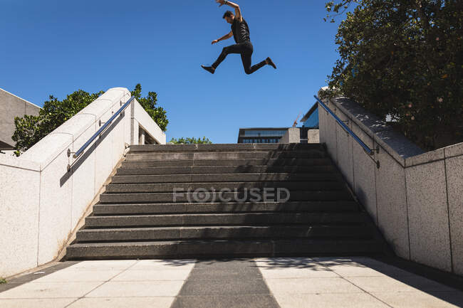 Seitenansicht eines kaukasischen Mannes, der an einem sonnigen Tag vor dem Gebäude in einer Stadt Parkour praktiziert und über Treppen springt. — Stockfoto