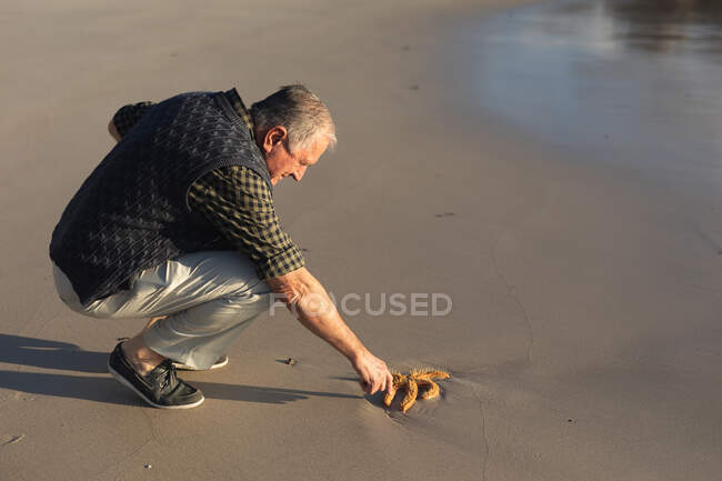 Vista lateral de un hombre caucásico mayor explorando solo en una playa, sentándose en cuclillas y extendiéndose para tocar una estrella de mar, varado en la arena - foto de stock