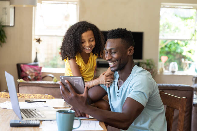 Chica afroamericana con una blusa amarilla, distanciamiento social en casa durante el bloqueo de cuarentena, pasar tiempo con su padre usando una tableta. - foto de stock