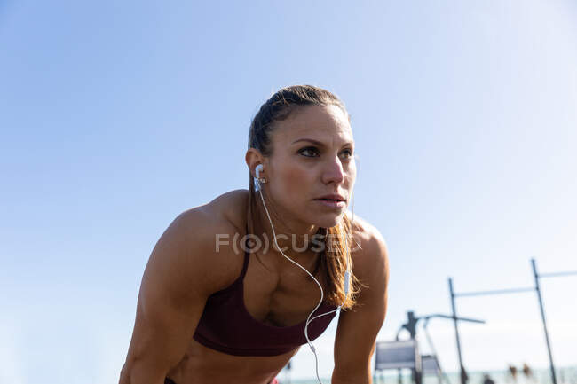 Low angle side view of a sporty Caucasian woman with long dark hair exercising in an outdoor gym during daytime, leaning on her knees with her headphones on. — Stock Photo