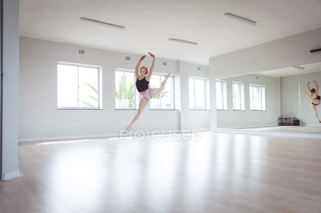 Caucasian attractive female ballet dancer with red hair dancing ballet, preparing for a ballet class in a bright studio, focusing on her exercise jumping in the air with her arms above her head. — Stock Photo