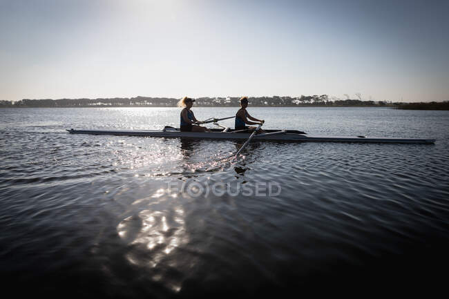 Vista lateral de dos remadoras caucásicas desde un equipo de remo entrenando en el río, remando en una concha de carreras en el agua, con la luz del sol reflejándose en las ondas en primer plano - foto de stock