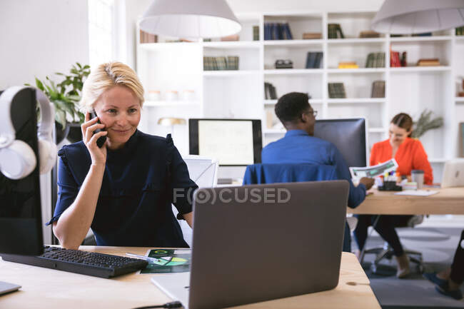 Улыбающаяся кавказская деловая женщина, работающая в современном офисе, сидящая за столом и за компьютером, разговаривающая на смартфоне, со своими деловыми коллегами, работающими на заднем плане — стоковое фото