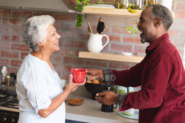 Счастливая пожилая афроамериканская пара дома, стоящая на кухне, разговаривающая, смеющаяся и пьющая кофе, дома вместе изолирующая во время пандемии коронавируса — стоковое фото