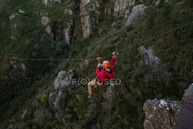Low angle side view of Caucasian man enjoying time in nature, zip lining on a cloudy day in mountains. Fun adventure vacation weekend. — Stock Photo