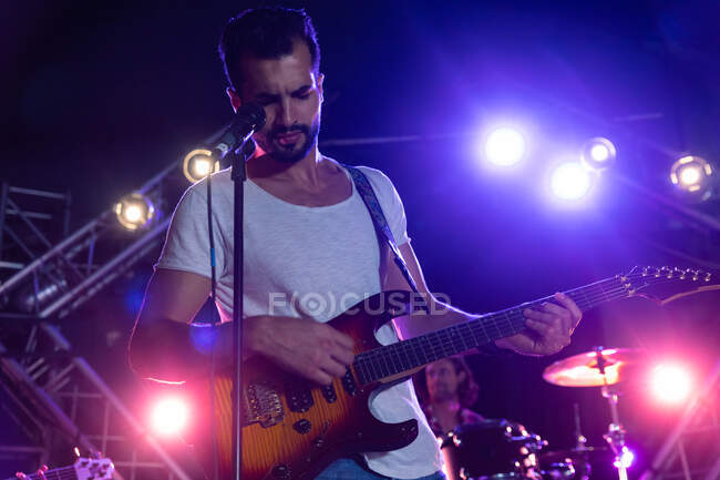 Vista frontal de un guitarrista caucásico centrado en tocar su guitarra eléctrica, de pie en un escenario iluminado con un micrófono en un soporte frente a él, actuando con una banda en un lugar de música - foto de stock