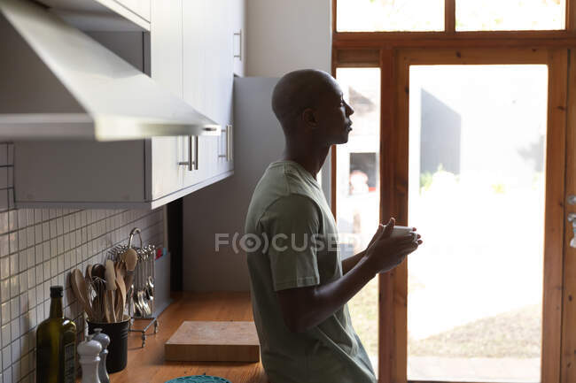 Vue latérale d'un Afro-Américain à la maison, debout dans la cuisine tenant une tasse de café et regardant loin — Photo de stock