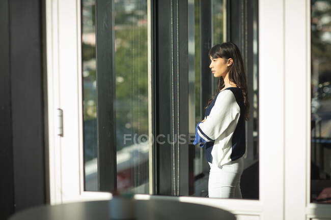 Una empresaria asiática que trabaja en una oficina moderna, mirando por una ventana y pensando, cruzando los brazos, en un día soleado - foto de stock