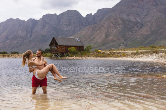Передній вид кавказької пари добре проводить час, подорожуючи горами, стоячи в озері, чоловік тримає жінку. — стокове фото