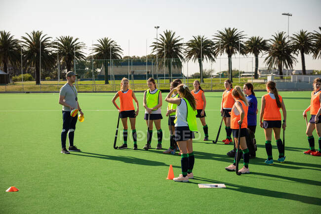 Side view of a group of female Caucasian field hockey players and their Caucasian male field hockey coach, training before a game, working out on a field hockey pitch, doing exercises, preparing for shooting training on a sunny day — Stock Photo