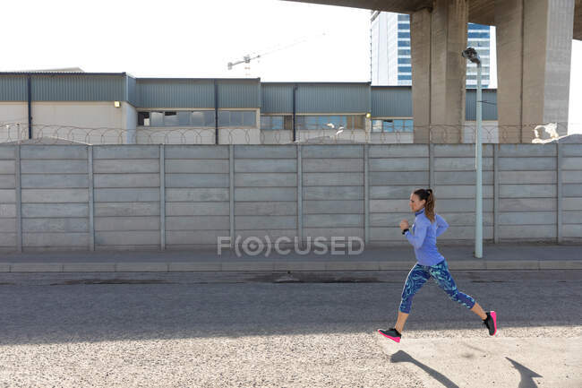 Боковой вид спортивной белой женщины с длинными темными волосами, тренирующейся в городской местности в солнечный день, бегущей. — стоковое фото