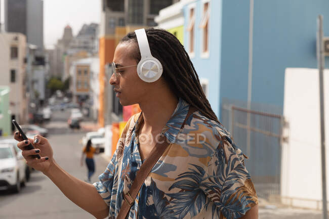 Vista lateral de cerca de un hombre de raza mixta con rastas largas en la ciudad en un día soleado, de pie en la calle con auriculares y usando un teléfono inteligente - foto de stock