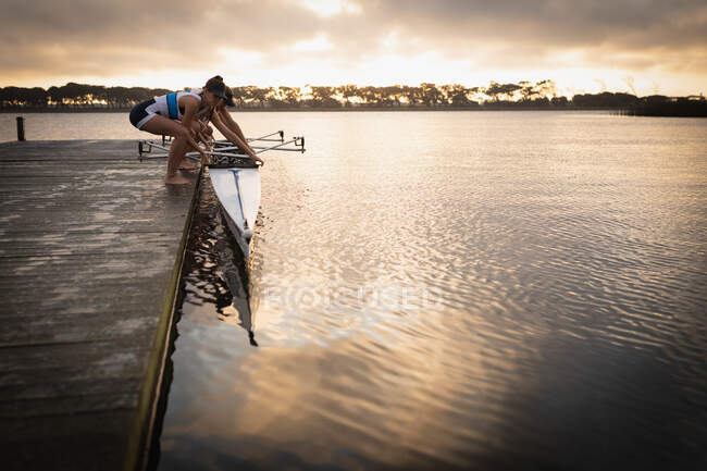 Vista laterale di una squadra di canottaggio di quattro donne caucasiche in piedi su un molo e mettendo una barca nel fiume in preparazione per l'allenamento di canottaggio — Foto stock