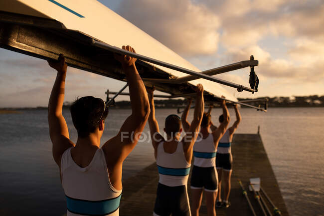 Visão traseira de uma equipe de remo de quatro homens caucasianos carregando um barco acima de suas cabeças com os braços levantados, andando ao longo de um molhe no rio ao pôr do sol — Fotografia de Stock