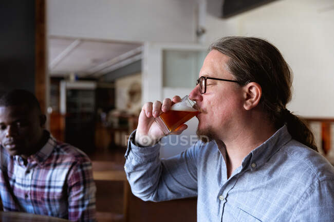 Kaukasischer Mann mit langen Haaren probiert Bier in einer Mikrobrauerei, im Hintergrund sitzt ein Afroamerikaner an einem Tisch. — Stockfoto