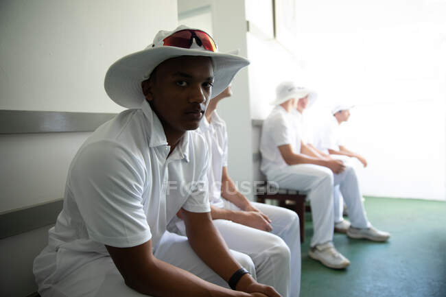 Передній вигляд закритий підлітка афроамериканського гравця в крикет у білому одязі, сидить на лавці в мінливій кімнаті і дивиться на камеру, а інші гравці сидять на задньому плані.. — стокове фото