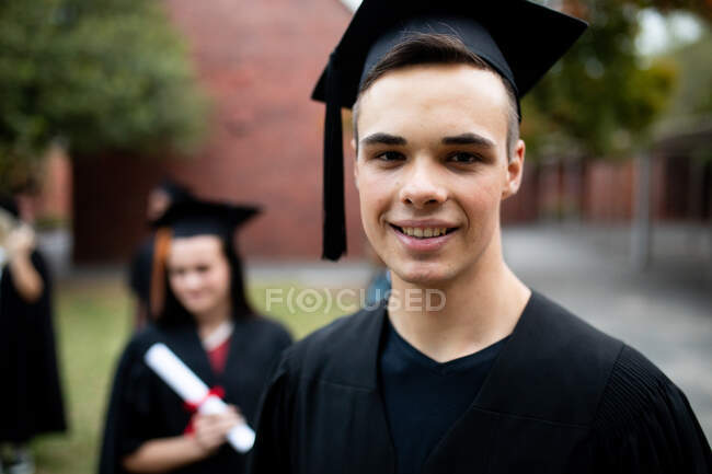 Retrato de un adolescente caucásico estudiante de secundaria con una gorra y un vestido en su día de graduación, mirando a la cámara y sonriendo, con otros estudiantes con gorras y batas en el fondo - foto de stock