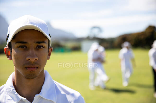 Portrait of a confident teenage Asian male cricket player wearing cricket whites and a cap, standing on a cricket pitch on a sunny day looking to camera, with other players standing in behind. — Stock Photo