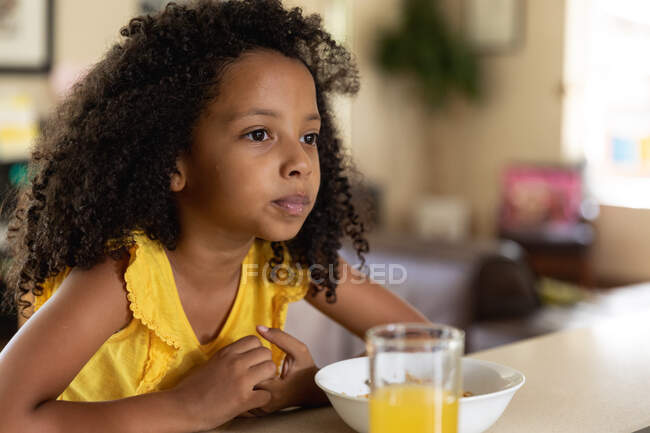 Chica afroamericana, distanciamiento social en casa durante el encierro de cuarentena, sentada junto a una mesa y desayunando y un vaso de jugo de naranja. - foto de stock