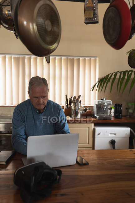 Front view of a senior Caucasian man relaxing at home, sitting at the counter in his kitchen using a laptop, with a VR headset sitting on the pan in front of him and pans and kitchen utensils hanging in the foreground — Stock Photo