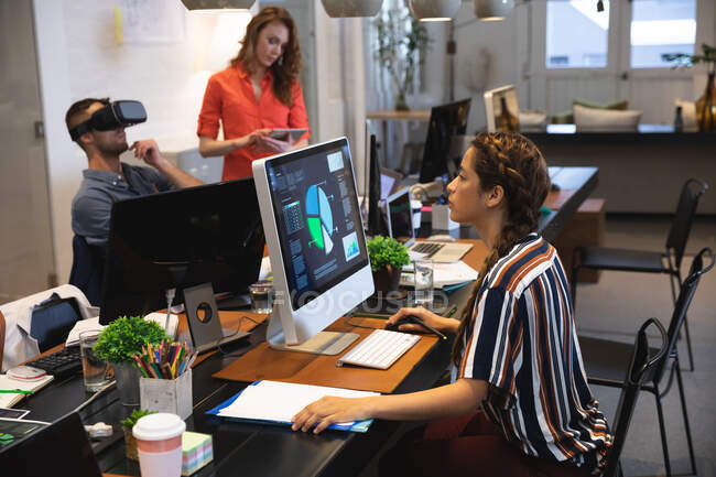 Raza mixta mujer de negocios creativo trabajando en una oficina moderna informal, sentado en una mesa y utilizando un ordenador con colegas que trabajan en el fondo - foto de stock