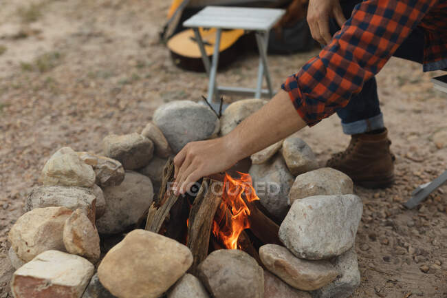 Side view close up of man having a good time on a trip to the mountains, squatting by a campfire, putting a log into a fire — Stock Photo