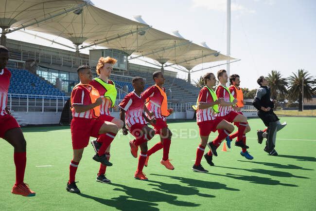 Side view of a multi-ethnic group of teenage male field hokey players, training with their coach before a game, working out on a field hockey pitch on a sunny day, running on the spot — Stock Photo